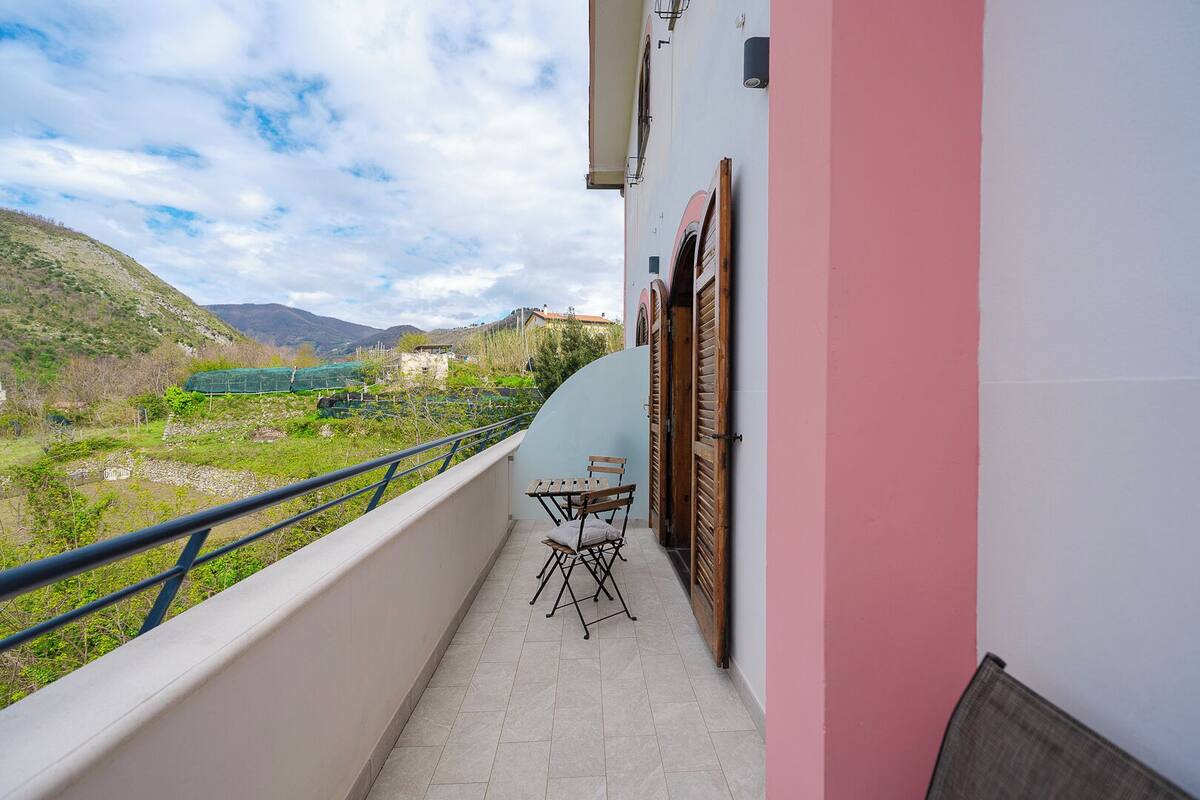 A balcony with a view of a green valley and mountains. There is a table and two chairs on the balcony.