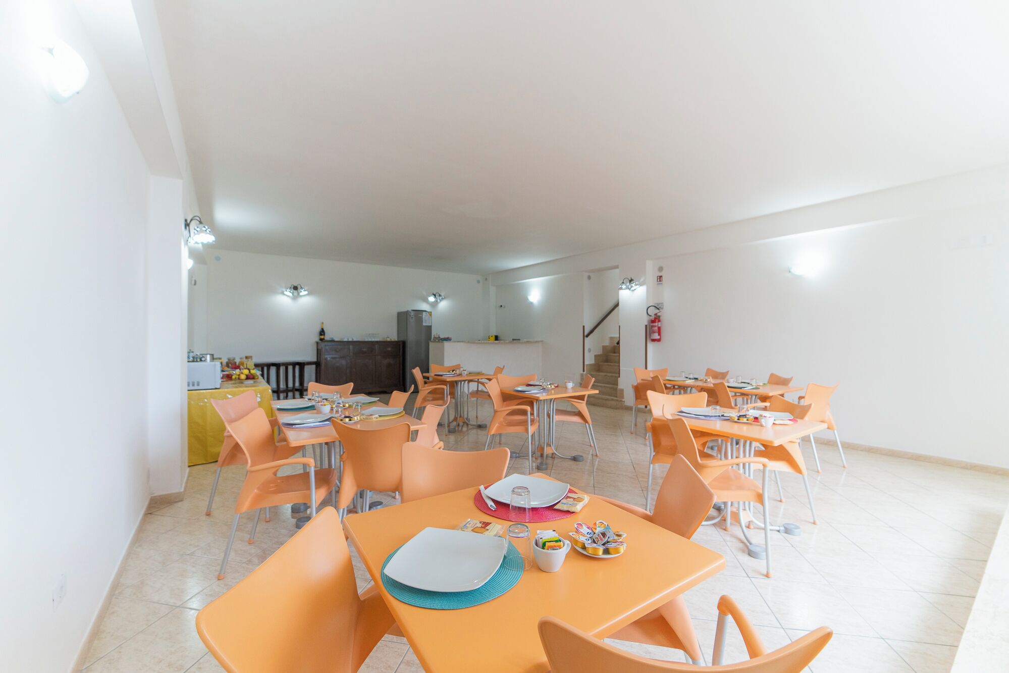 Un comedor amplio con varias mesas y sillas, una cocina al fondo y una escalera que sube a un nivel superior.