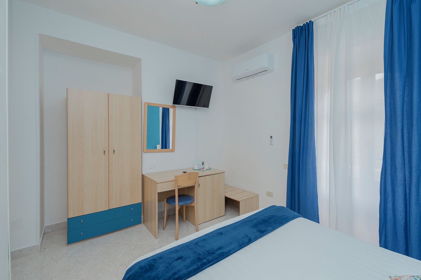 Una camera da letto con un letto, una scrivania, una sedia, un armadio, una TV e una finestra con tende.