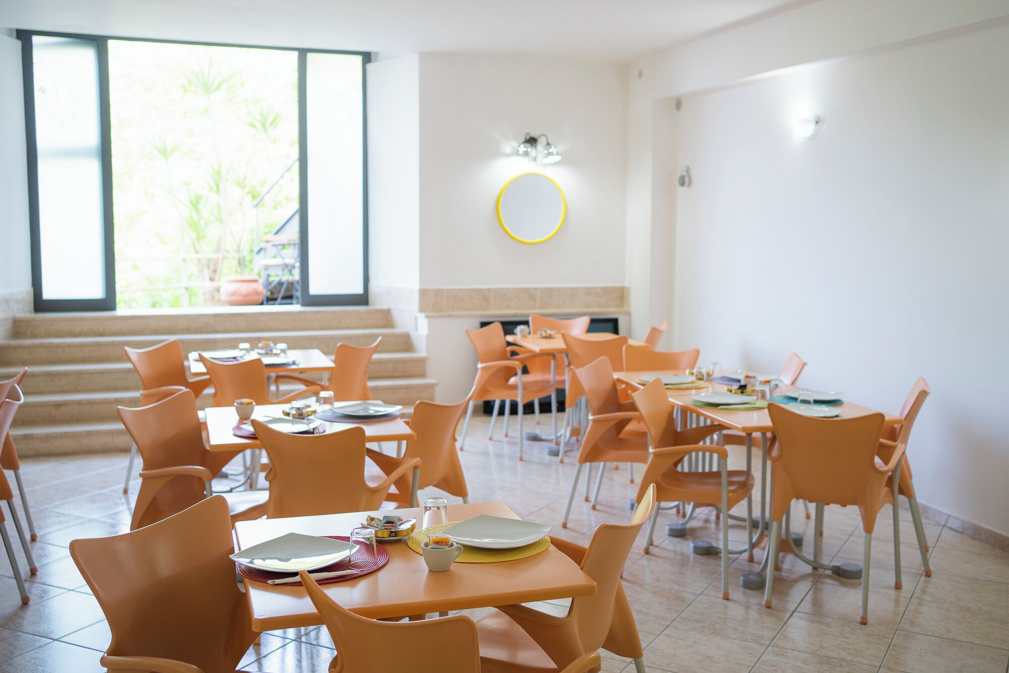 Un comedor luminoso y moderno con sillas y mesas naranjas, ventanas grandes y un espejo circular en la pared.