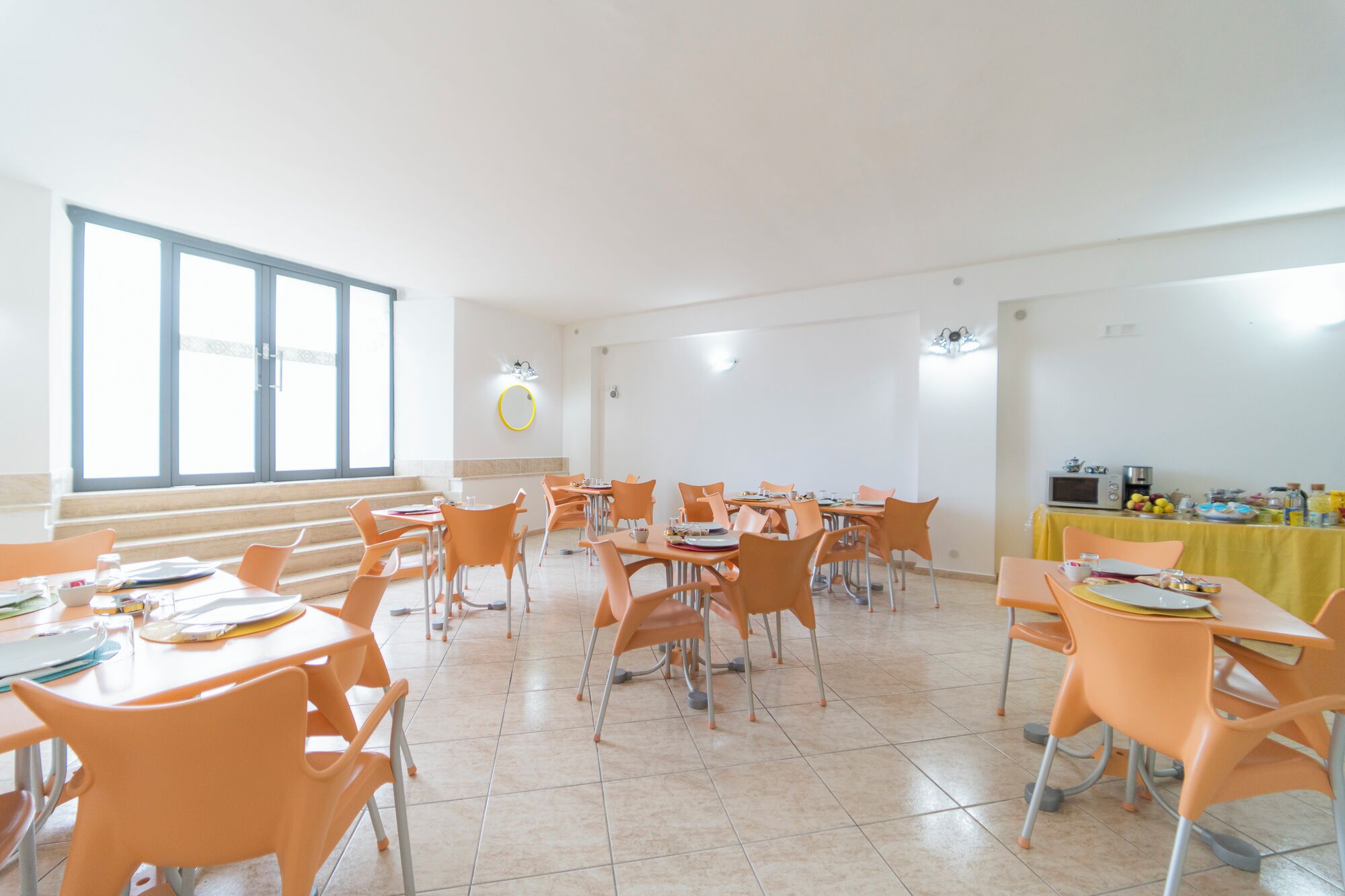 Un comedor amplio con sillas anaranjadas y mesas, ventanas grandes y una mesa cubierta con mantel amarillo, con comida y bebidas.