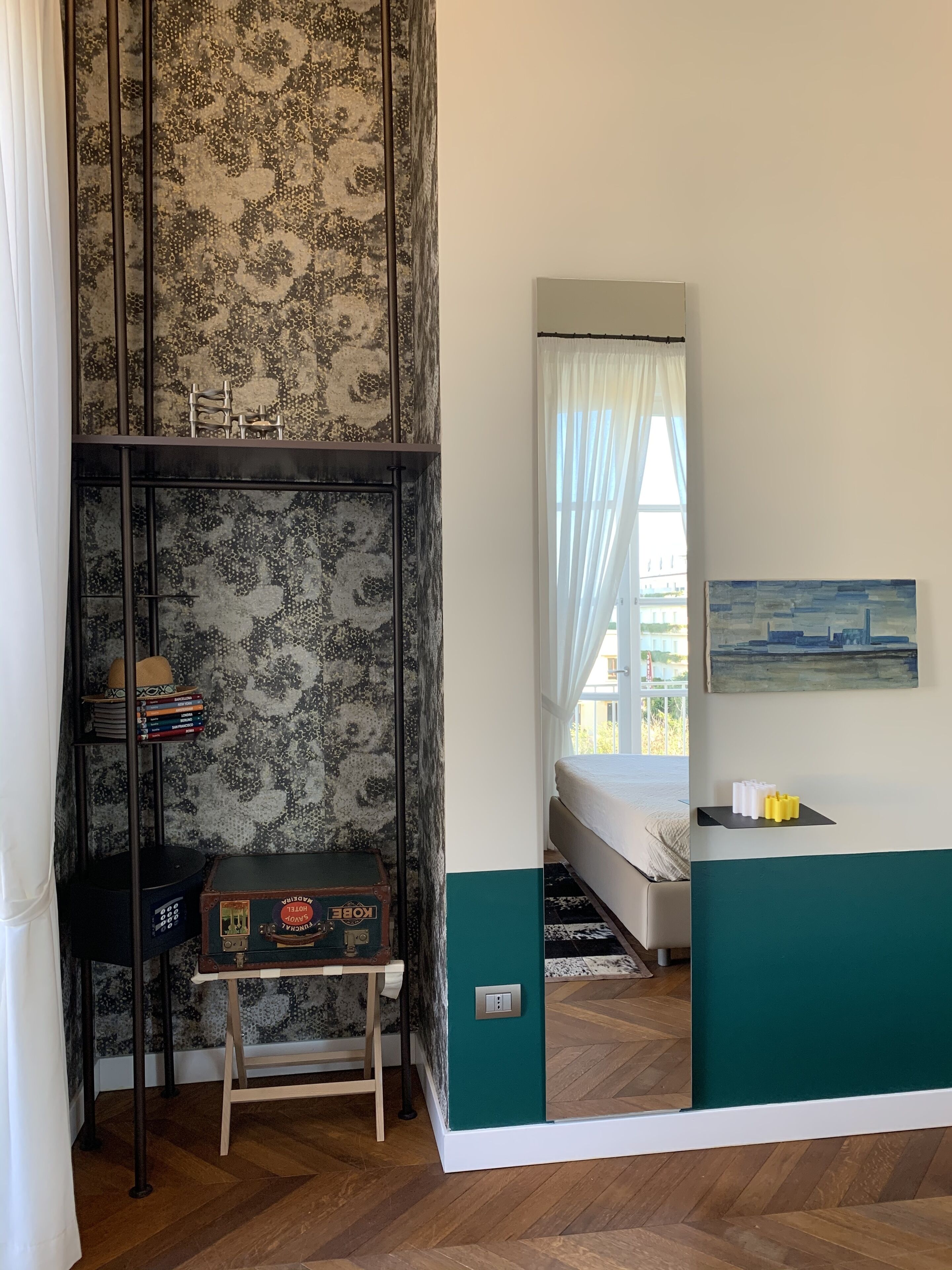 A bedroom with a bed, a desk with a suitcase, a bookshelf, and a painting on the wall.
