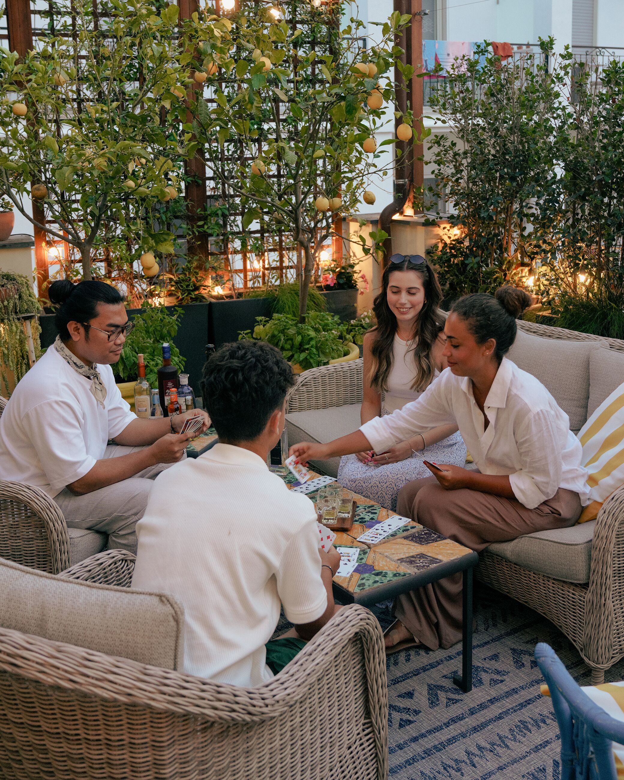 A group of people playing cards at an outdoor table with wicker furniture and a decorative tablecloth.