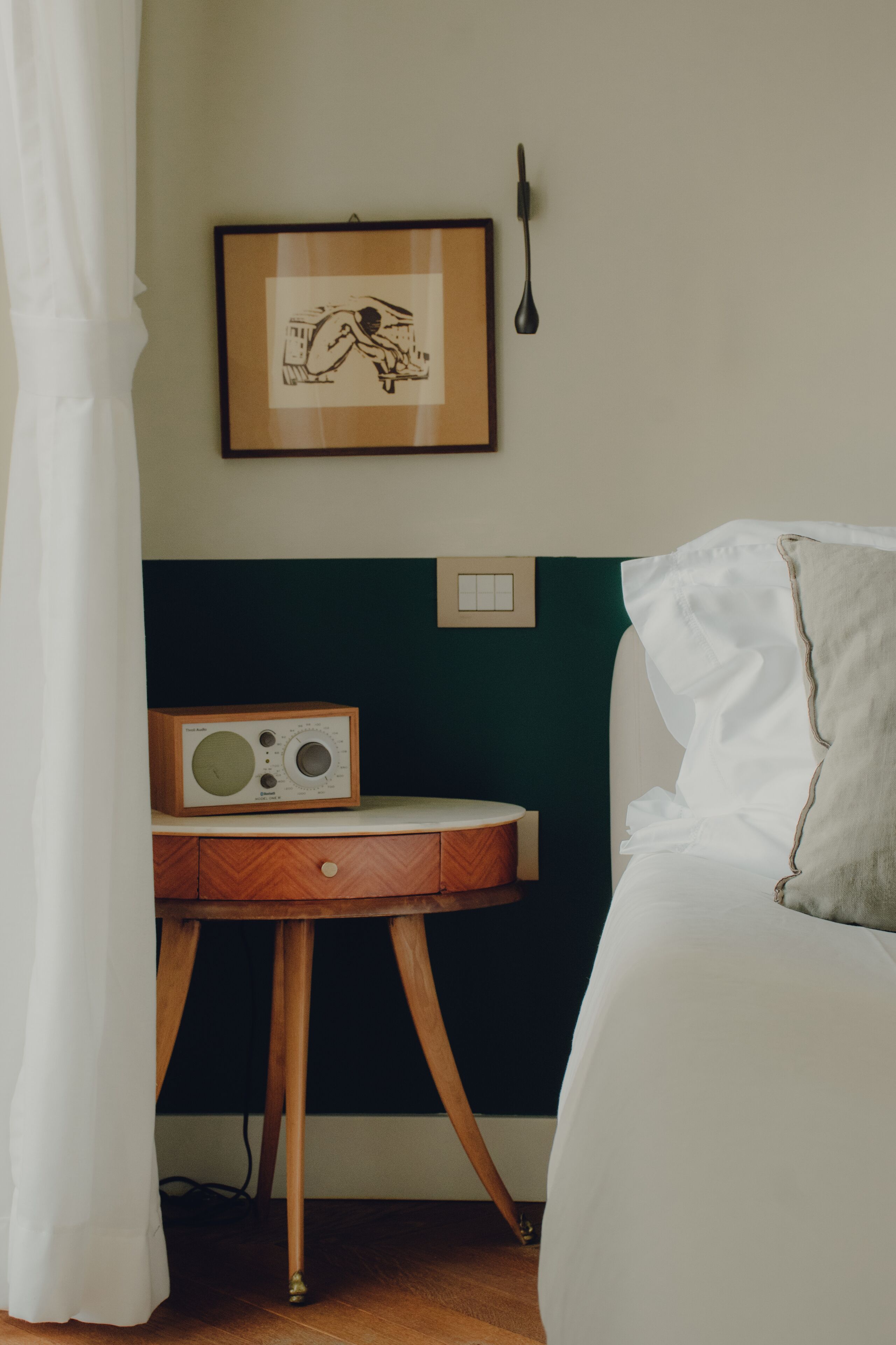 A bedroom with a bed, a bedside table with a radio, a framed picture on the wall, and a curtain.