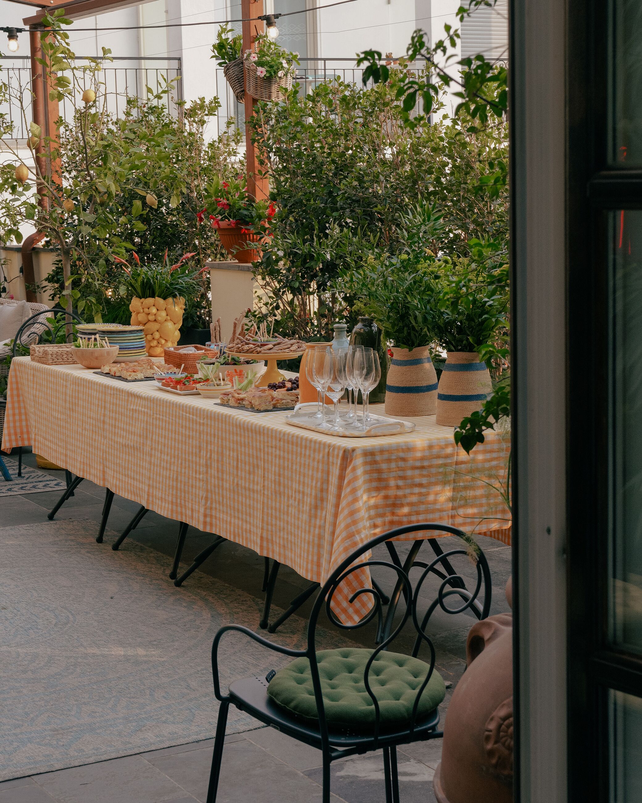 Un área de comedor al aire libre con mantel a cuadros, diferentes alimentos y plantas en macetas.