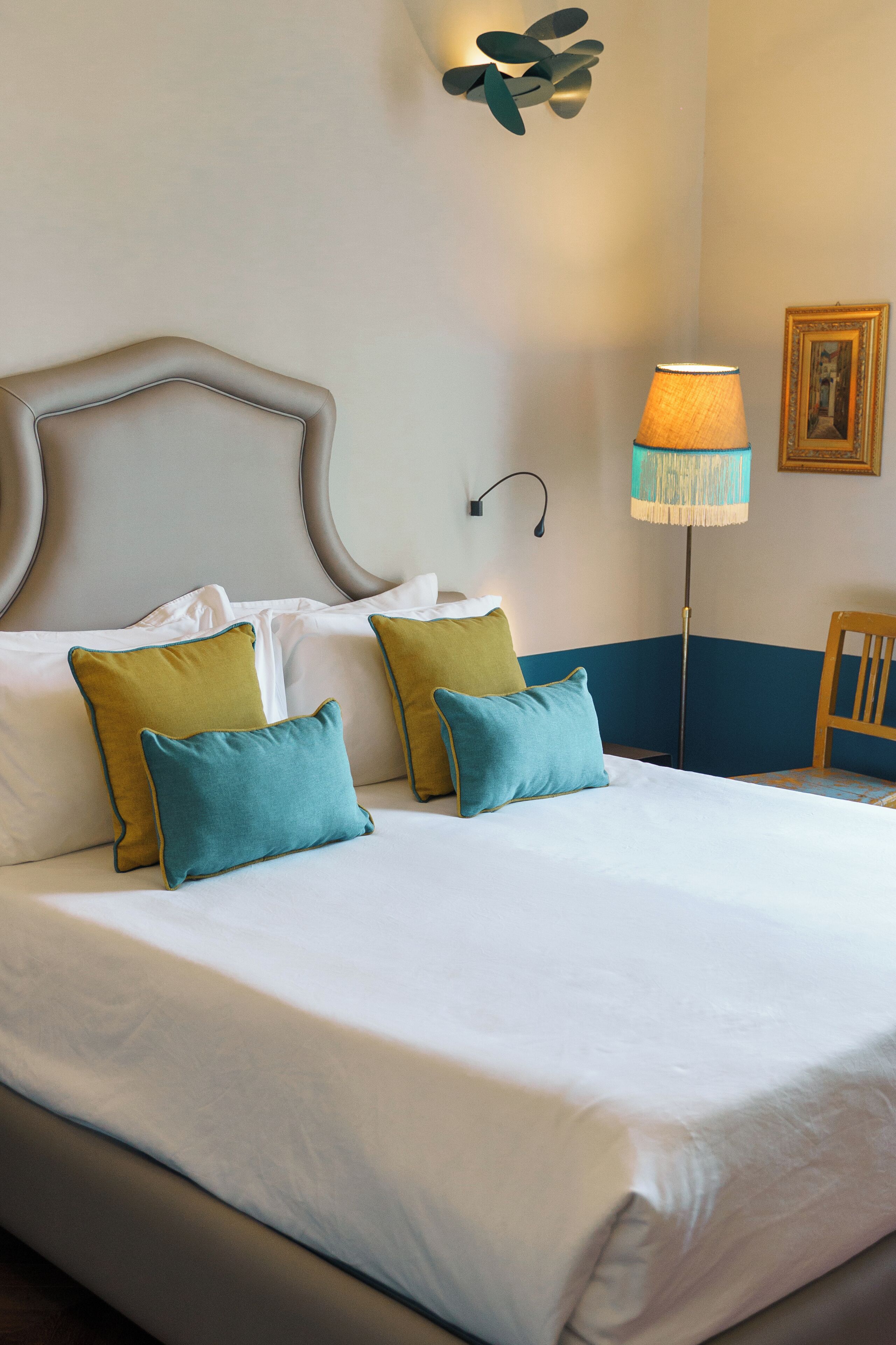Cama bem arrumada com lençóis brancos e almofadas verde-água e mostarda, abajur com cúpula azul e branca, e quadro na parede.
