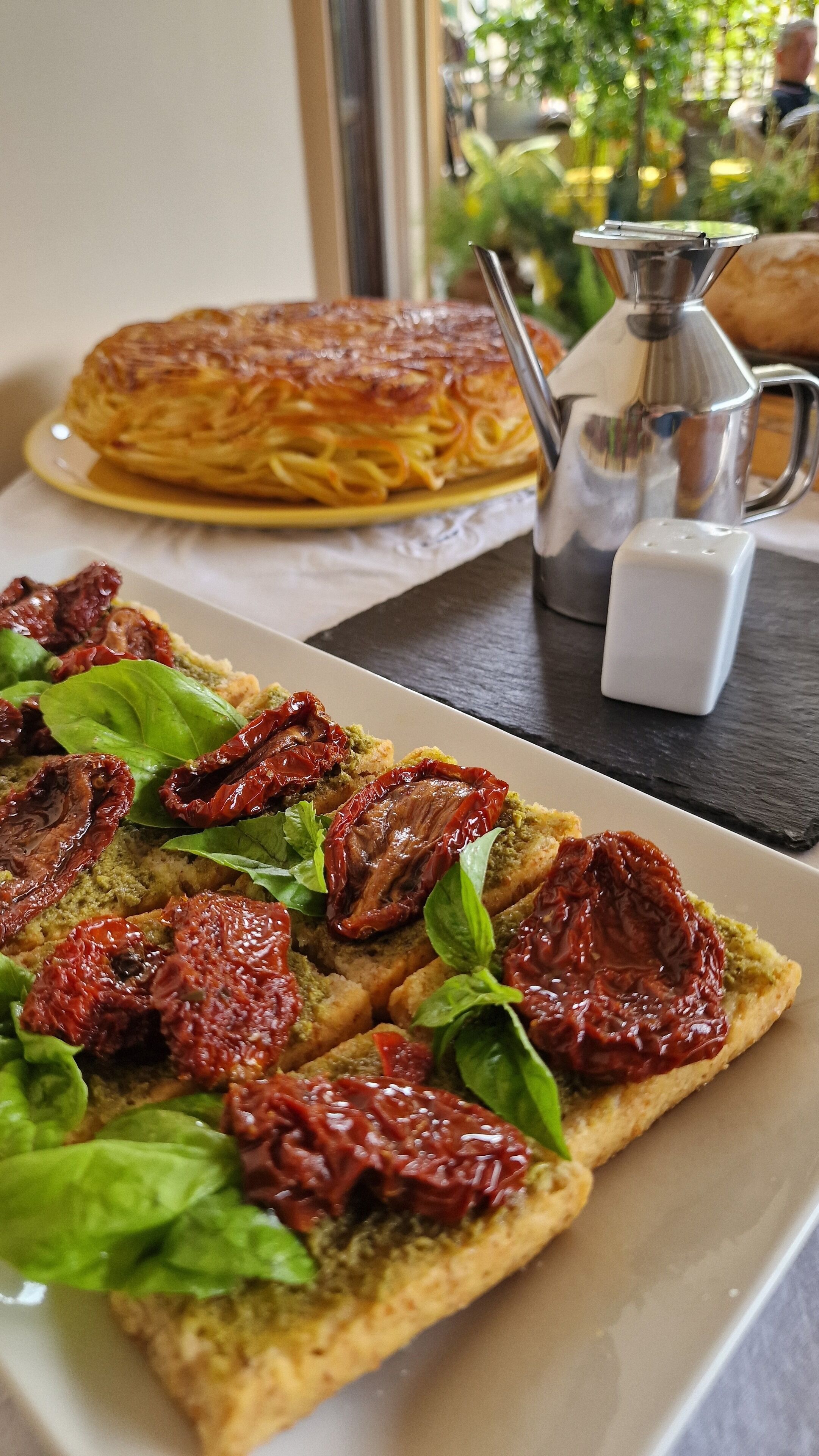 A plate of pizza with sun-dried tomatoes and basil, a glass of coffee, and a pastry on a table.