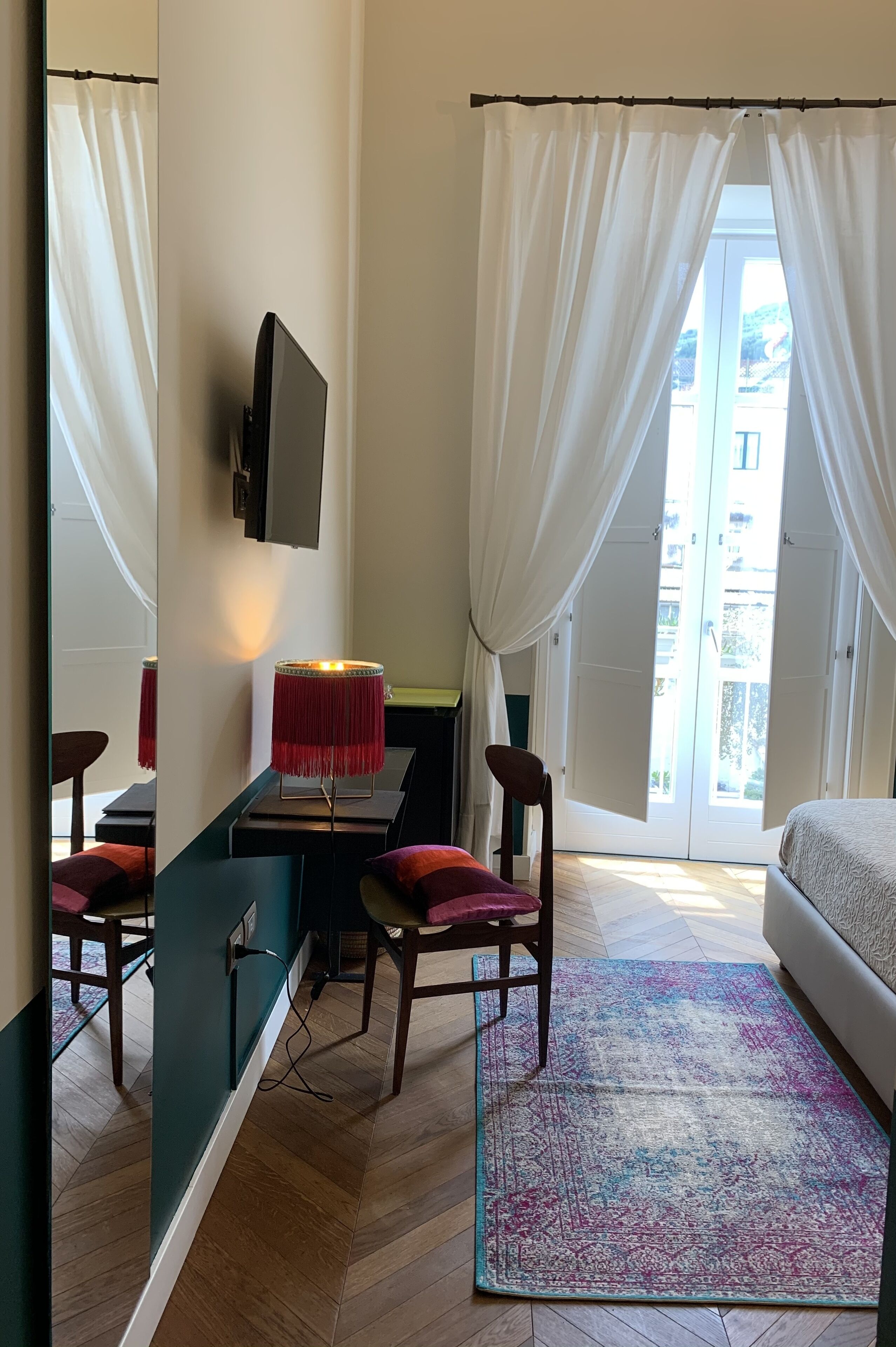 A hotel room with a bed, a desk with a chair, a TV mounted on the wall, and a view of the outside through a large window with curtains.