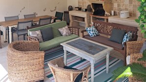 Un espace extérieur accueillant avec des meubles en osier, une cheminée en pierre et un panneau indiquant la « zone barbecue ».