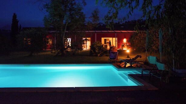 A swimming pool with lounge chairs, a house with a red exterior, and a person working near the house at night. - BRIGHT AND REFINED VILLA WITH POOL IMMERSED IN THE GREEN OF THE HILLS OF LUCCA (LUCCA)