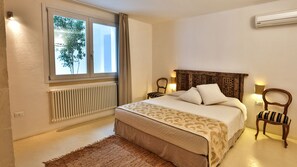 Ein Schlafzimmer mit Bett, Vorhängen am Fenster, Heizkörper, einem Holz-Kopfende, einem Nachttisch und einem Sessel.