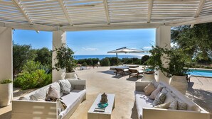 A covered outdoor seating area with a pool and lounge chairs, surrounded by greenery and a clear view of the sea.