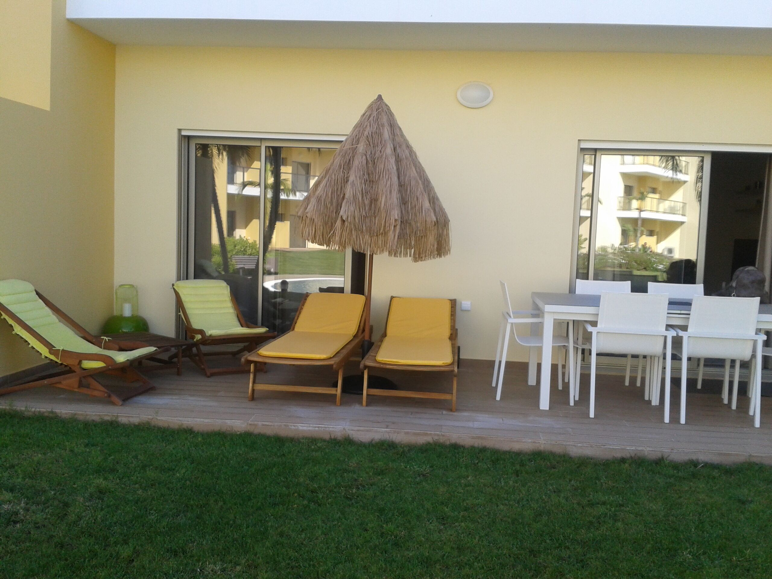 Une terrasse avec des chaises longues et une table, un parasol en chaume, et une vue sur un bâtiment à travers de grandes fenêtres.