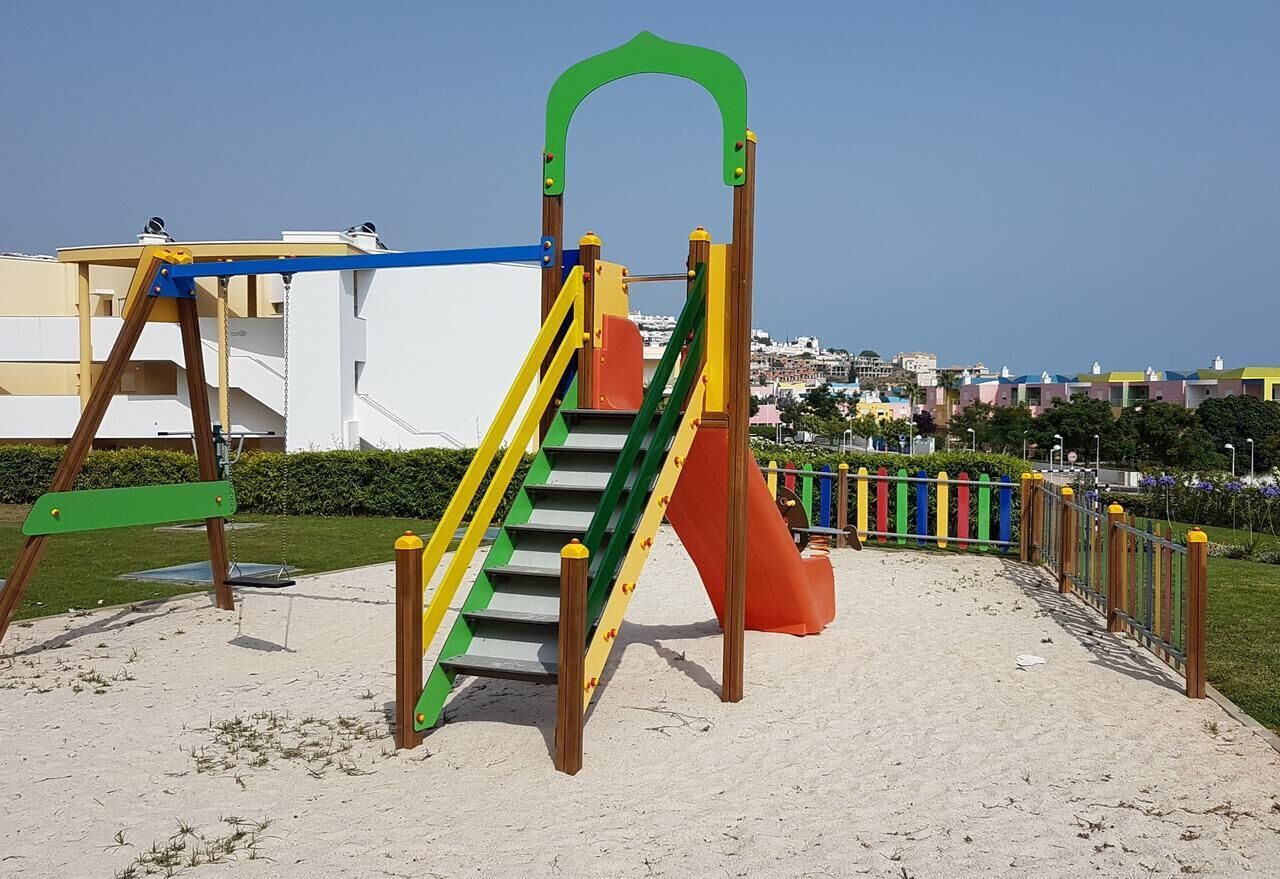 Une aire de jeux avec un toboggan et une structure d’escalade, entourée d’une zone sablonneuse et d’une clôture.