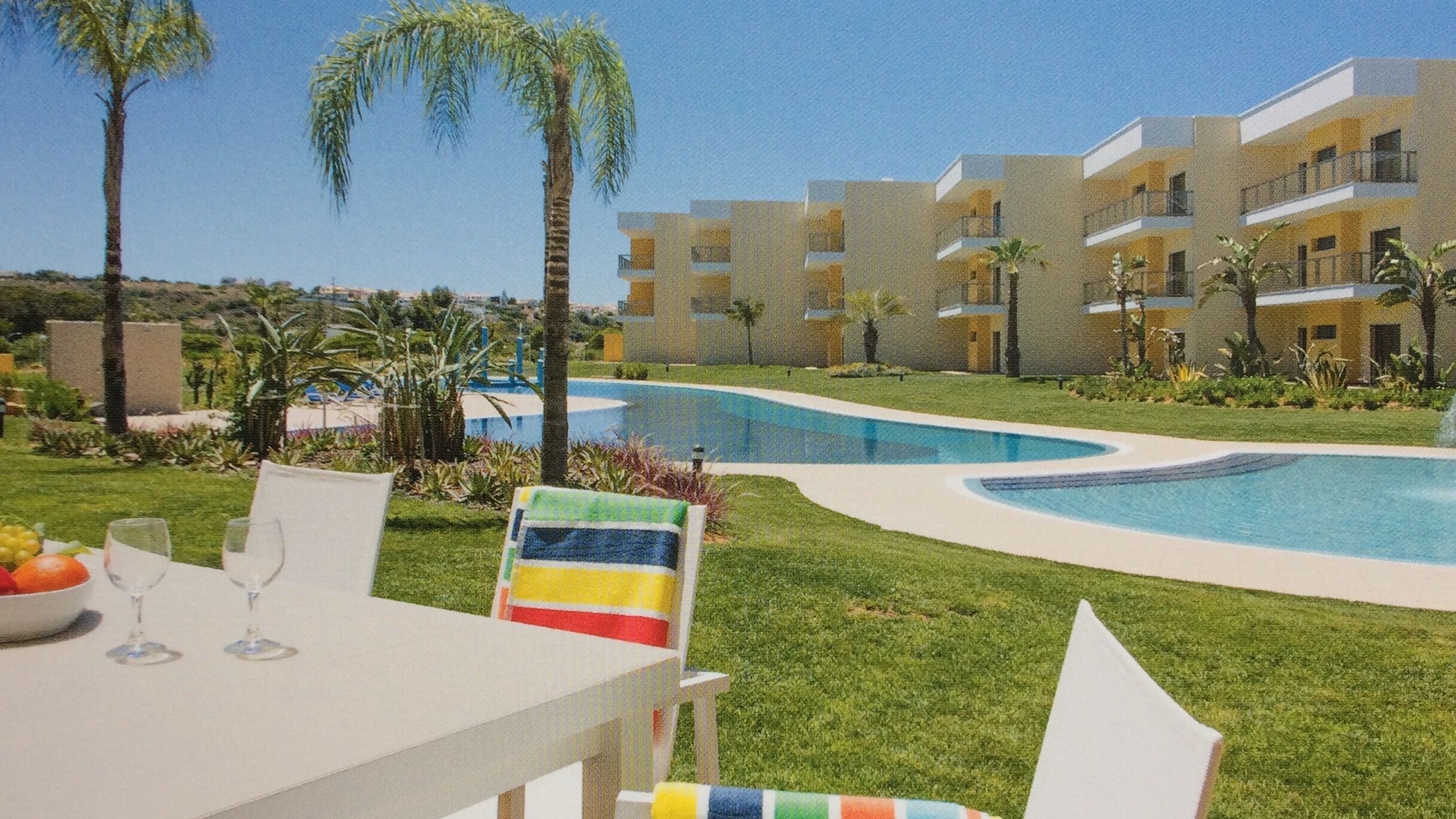 A pool area with a curved design, surrounded by greenery and palm trees. There are multiple-story buildings in the background.