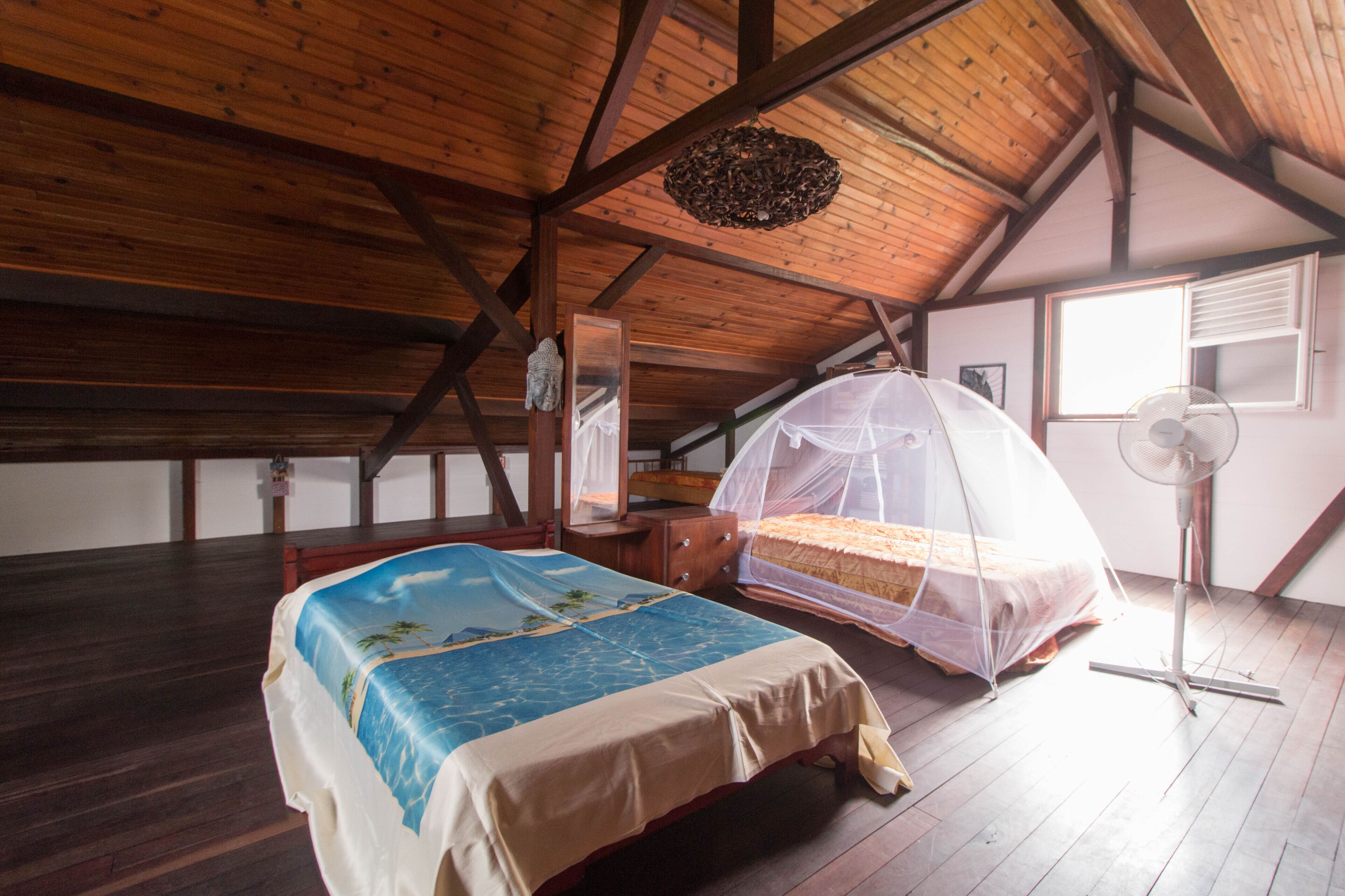 Een slaapkamer met houten plafond, een bed met klamboe, nachtkastje en een staande ventilator.