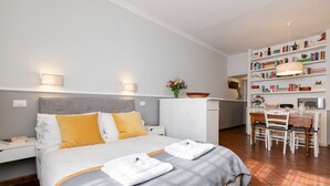 A neatly arranged hotel room with a bed, bedside tables, a desk with chairs, a bookshelf, and a small table with a vase of flowers. - Paradiso | Romantic open space in the heart of Rome (Roma)