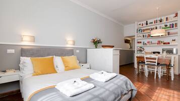 Une chambre d’hôtel bien rangée, avec un lit, des tables de chevet, un bureau avec des chaises, une bibliothèque et une petite table sur laquelle est posé un vase de fleurs.