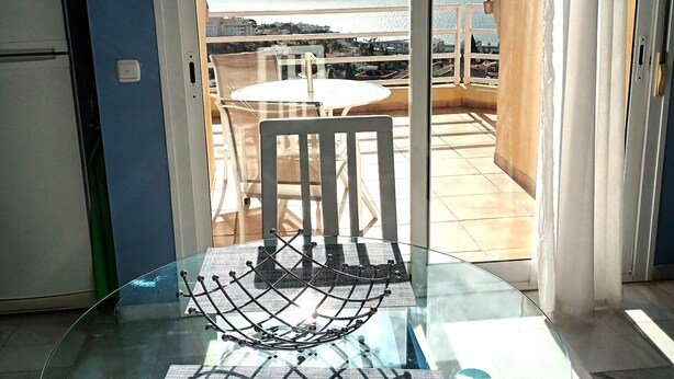 A glass-top dining table with a chair, a white door leading to a balcony with a view of the sea, and a refrigerator in the room.