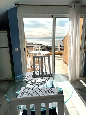 Een glazen eettafel met een stoel, een witte deur naar een balkon met uitzicht op zee, en een koelkast in de kamer.