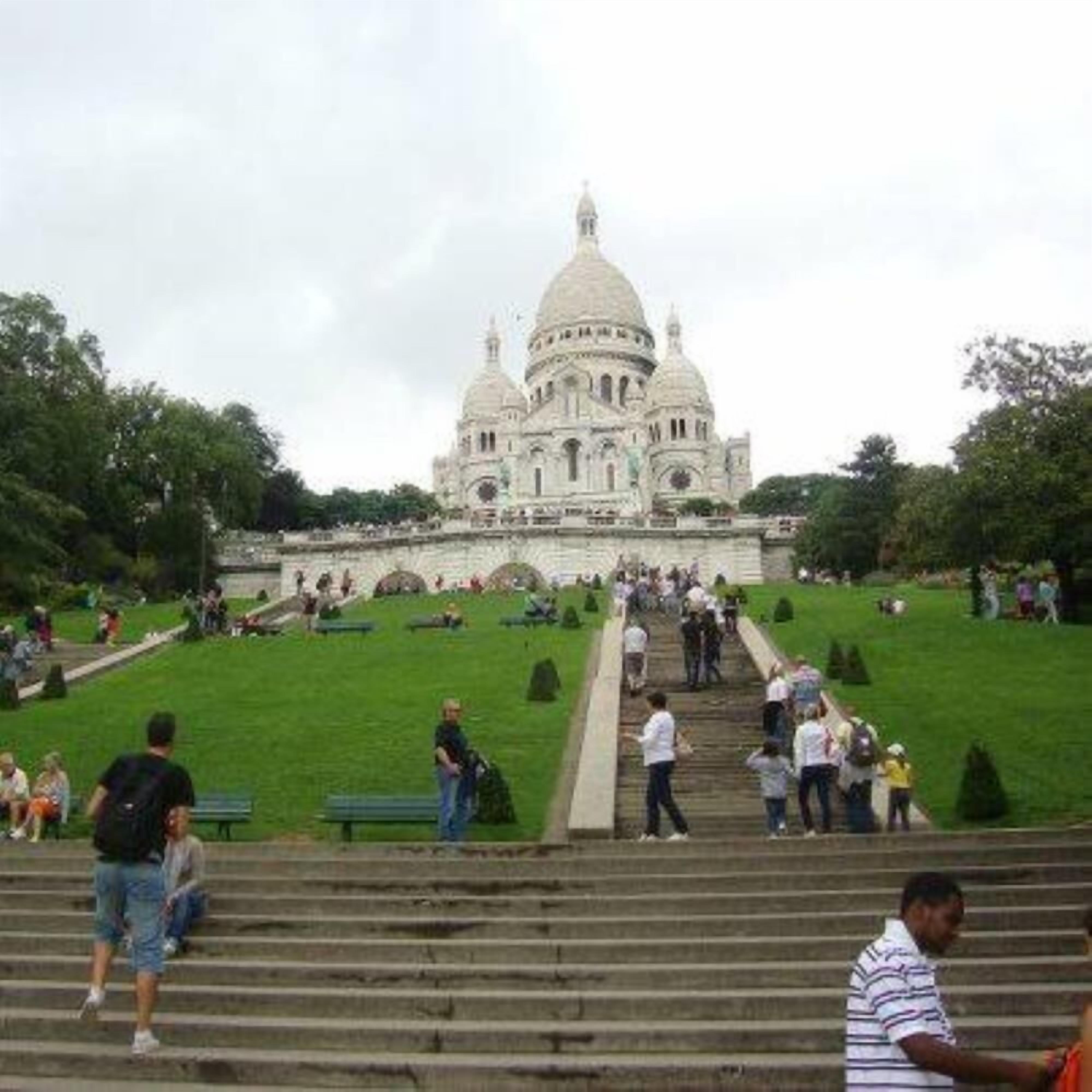 Un grand bâtiment en forme de dôme, avec des marches menant à l’entrée, entouré d’un parc verdoyant où l’on trouve des bancs et des passants.