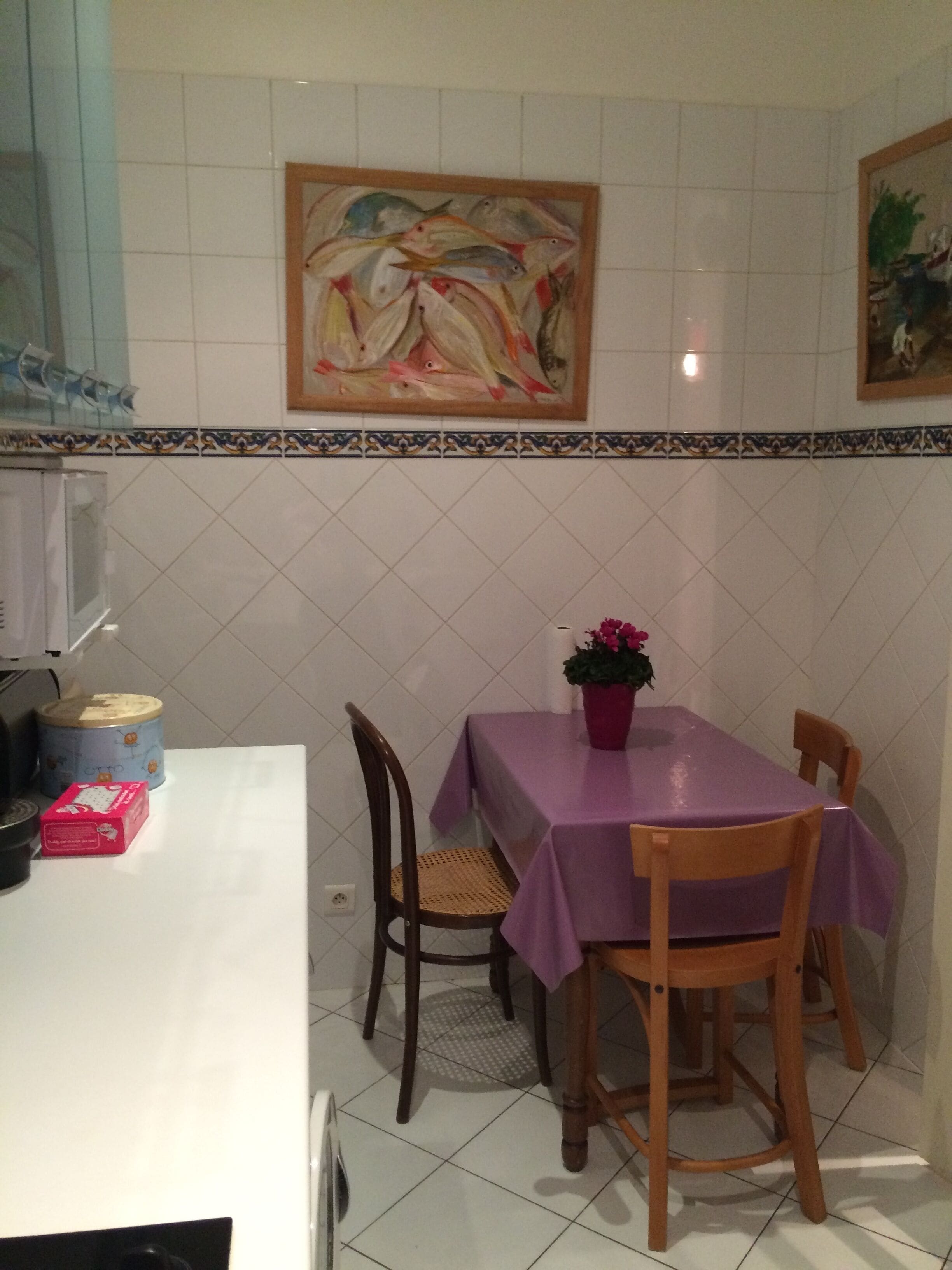 Un comedor con una mesa cubierta con mantel morado, rodeada de sillas de madera. Hay una planta en maceta con flores rosas sobre la mesa.
