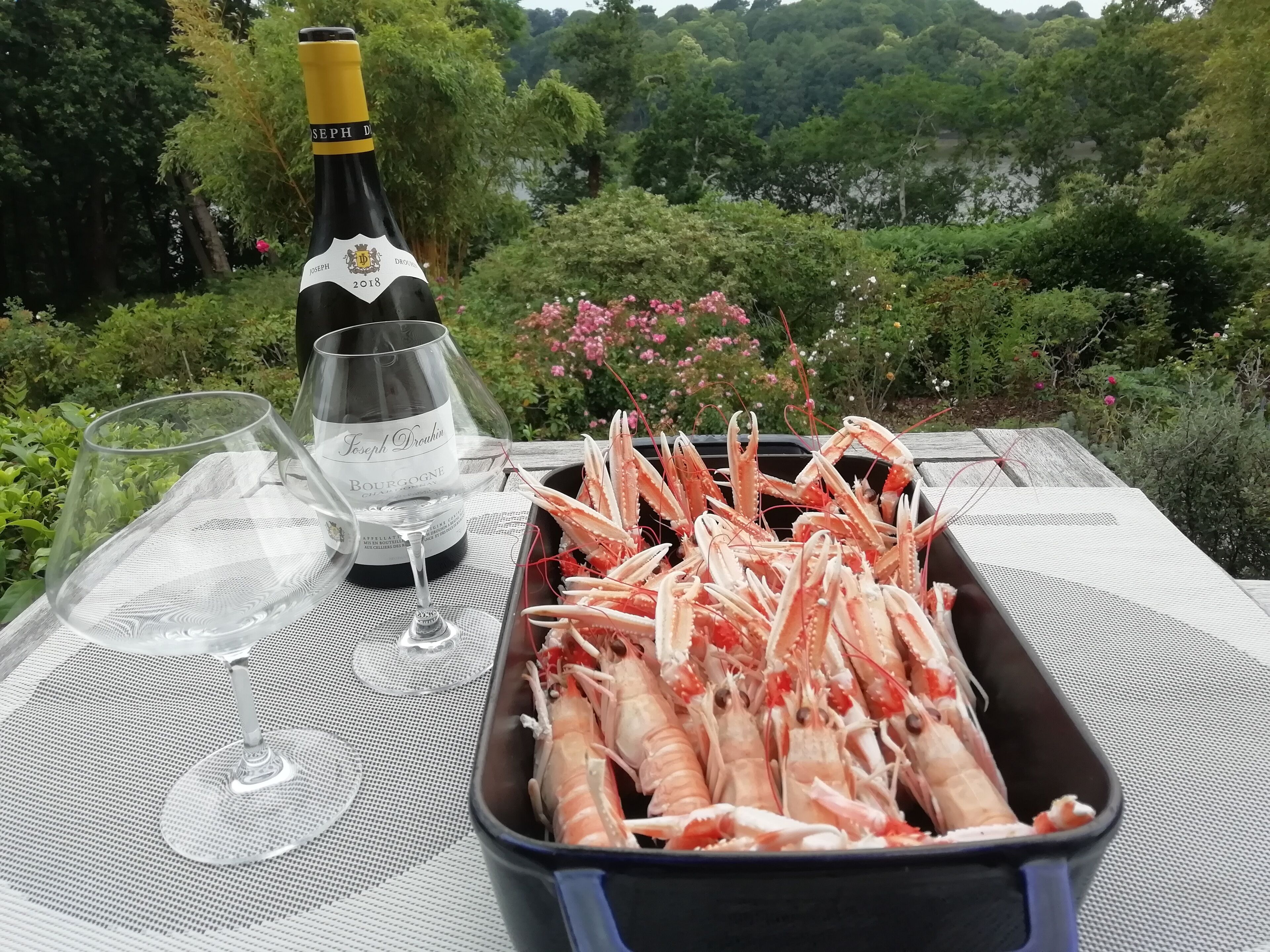 A bottle of Joseph Drouhin wine, a glass, and a tray of cooked crayfish on an outdoor table with a scenic view.