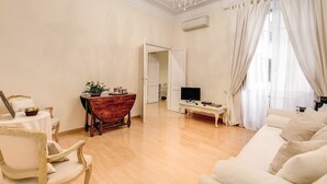 Amplia sala de estar con piso de madera, un sofá blanco, una mesa de centro de madera, un televisor y una araña.