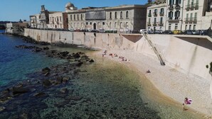 A coastal area with a rocky beach, clear water, and a large building with multiple windows and balconies. - Apartment in Ortigia furnished and equipped with all comforts (Siracusa)