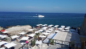 A seaside restaurant with outdoor seating overlooking the ocean, featuring a rocky coastline and a view of boats on the water. - Apartment in Ortigia furnished and equipped with all comforts (Siracusa)