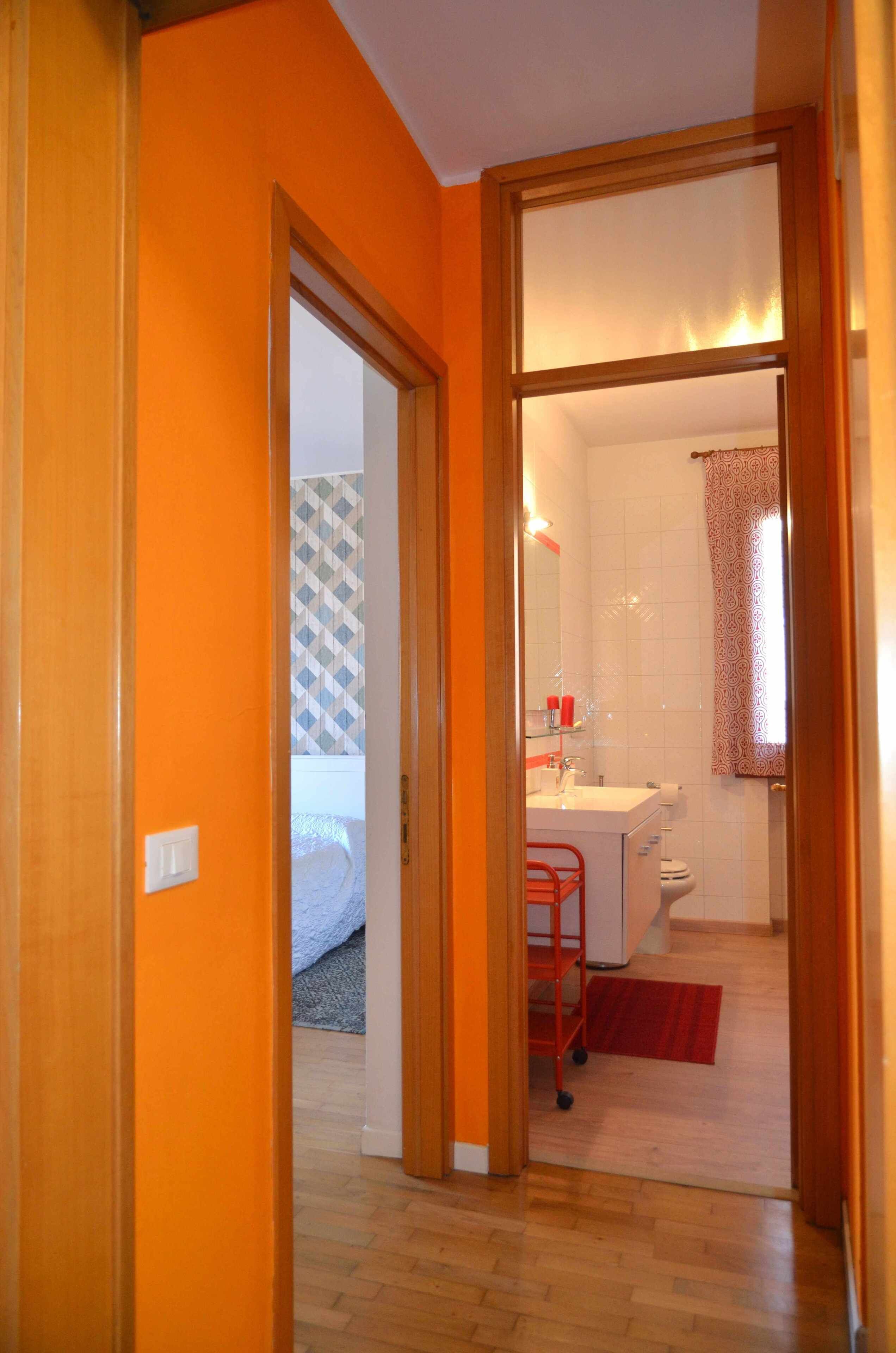 Et værelse med en orange væg, en trædør, en seng, et badeværelse med en hvid vask og en rød stol.