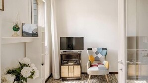 Ein Zimmer mit Fernseher auf einem Schrank, einem gemusterten Sessel, einer Vase mit Blumen und einem Regal mit Dekoration.