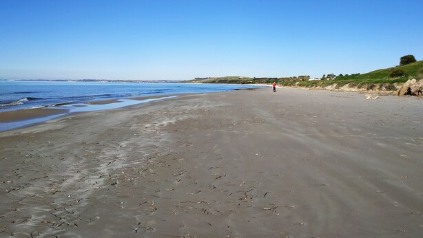 Een breed zandstrand met rustige golven, een helderblauwe lucht, en een persoon in het rood die van de camera afloopt.