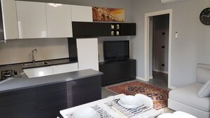 A modern kitchen with a built-in stove, a dining area with a table set for two, and a living area with a sofa and a TV.