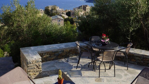 Un área de estar al aire libre con suelo de piedra, una mesa con sillas y vistas al mar y edificios a lo lejos.