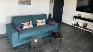 Un sofá color turquesa con almohadones estampados de pájaros, una mesita redonda con una vela y un televisor de pantalla plana sobre un soporte negro.