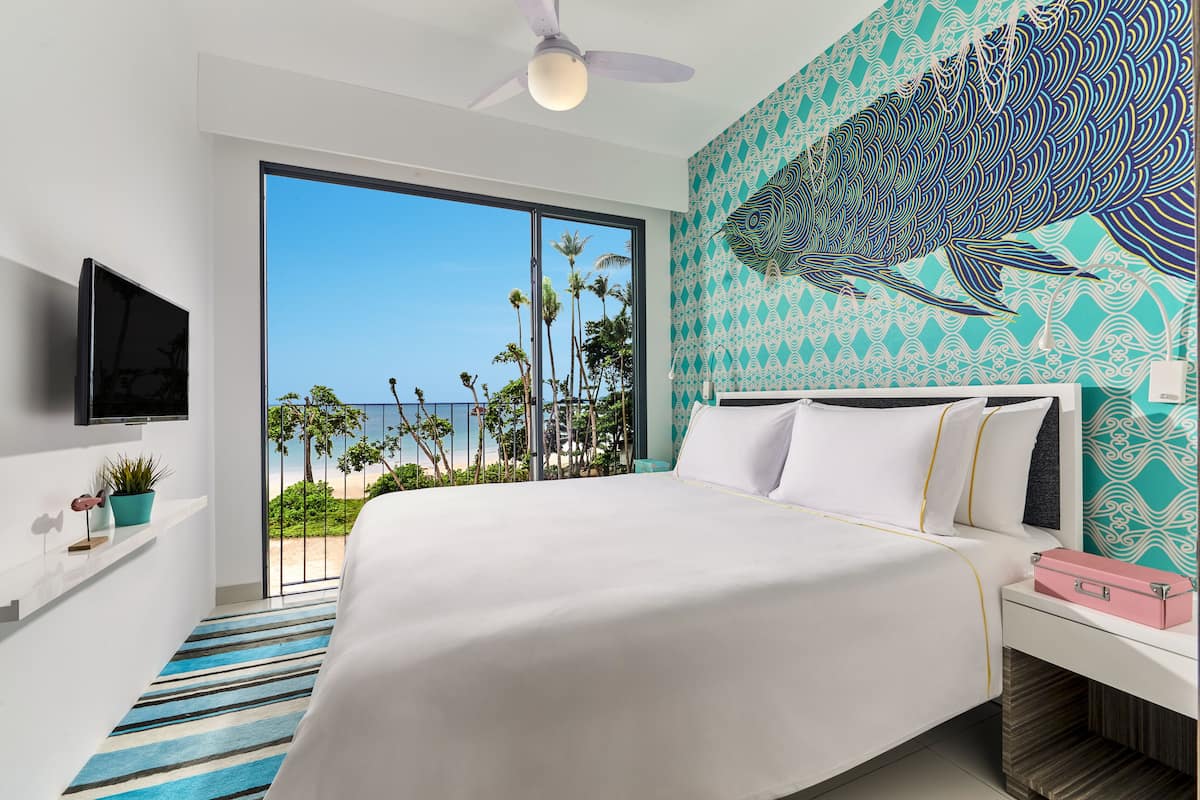A bedroom with a large bed, a TV mounted on the wall, a balcony with a view of palm trees and the ocean, and a decorative fish mural on the wall.