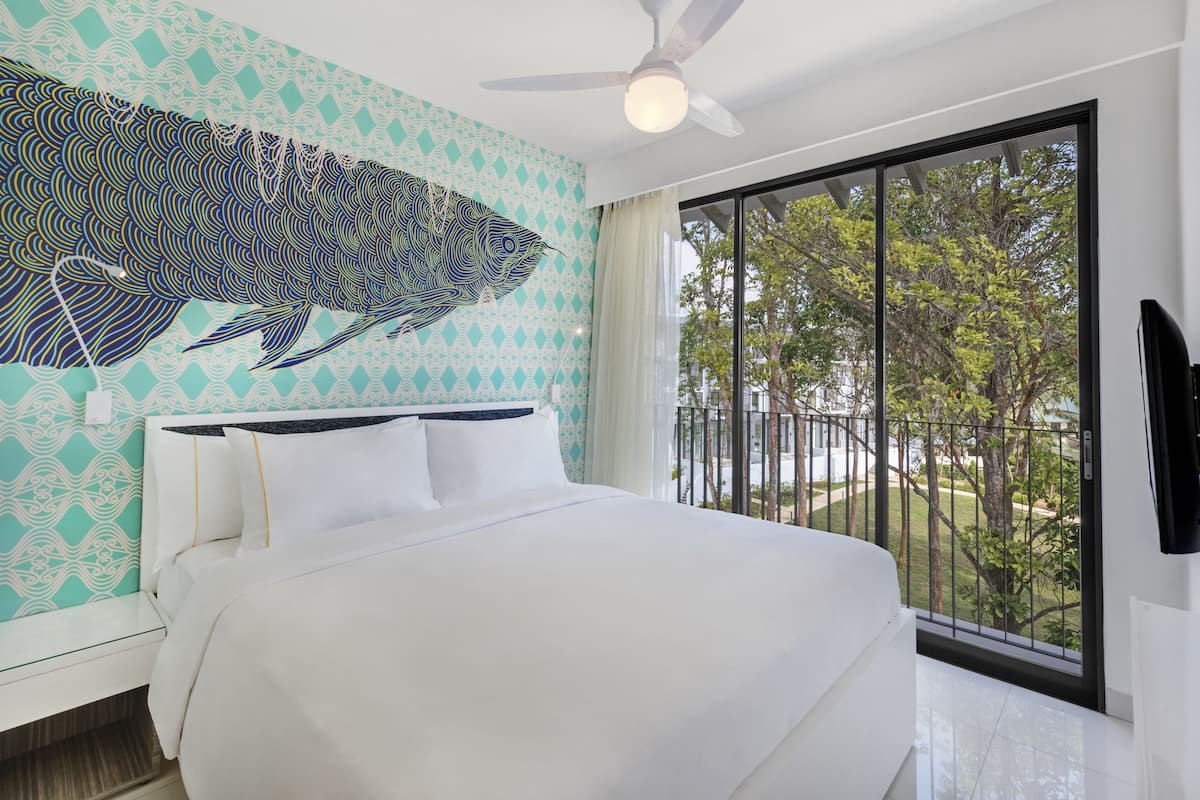 A modern hotel room with a large bed, a ceiling fan, and a view of the outdoors through a large window.