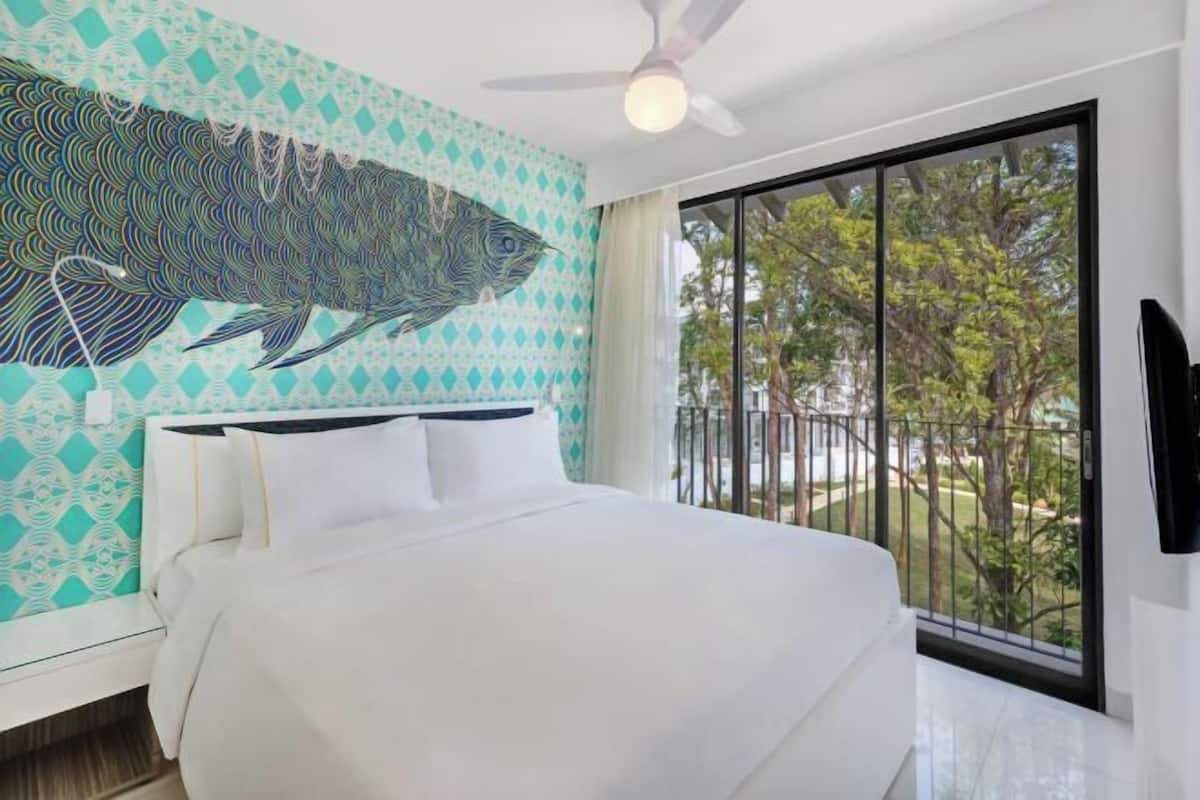 A hotel room with a bed, a ceiling fan, a large window with a view of trees, and a wall mural.