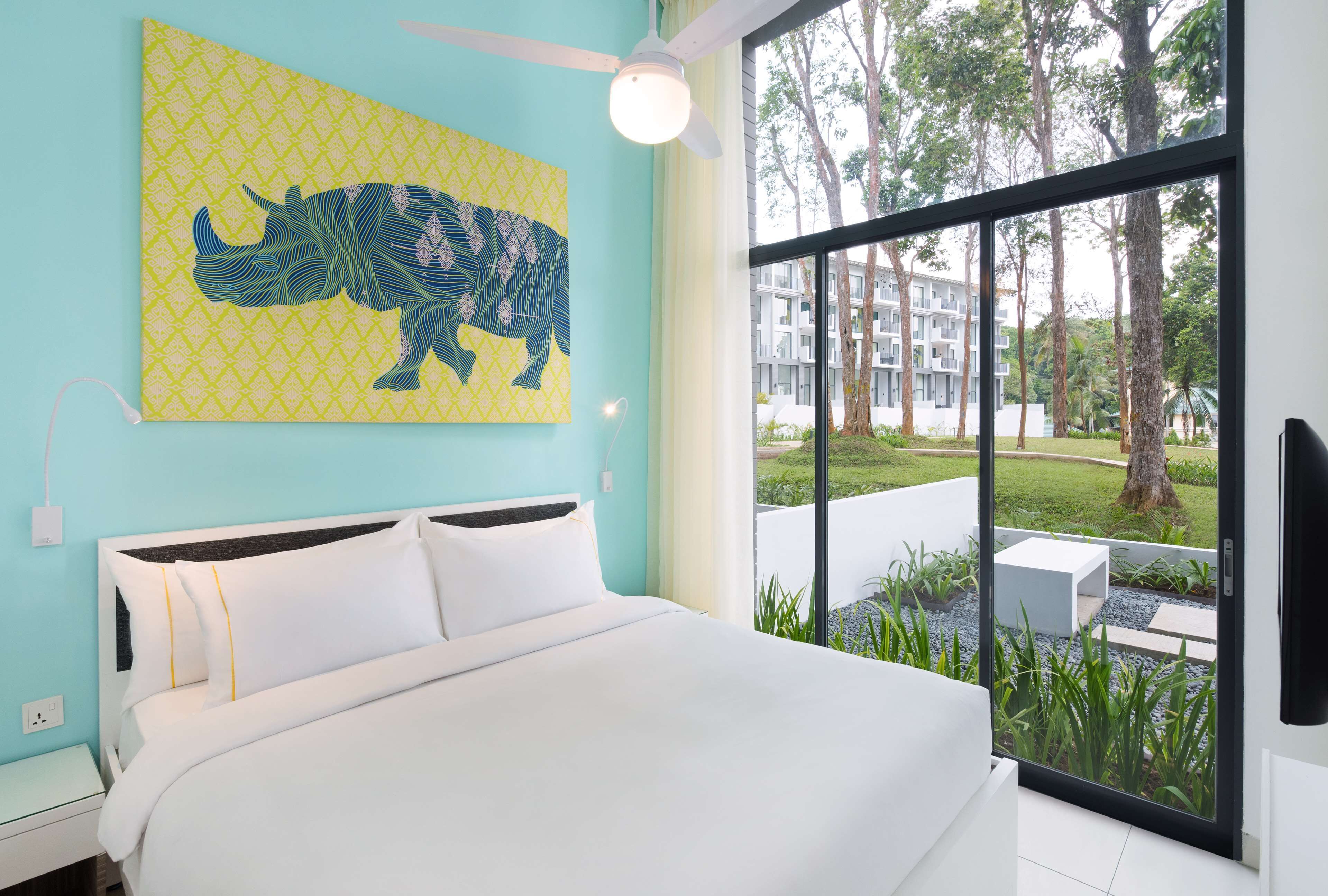A bedroom with a bed, a ceiling fan, a window with a view of a building and trees, and a wall with a geometric pattern.