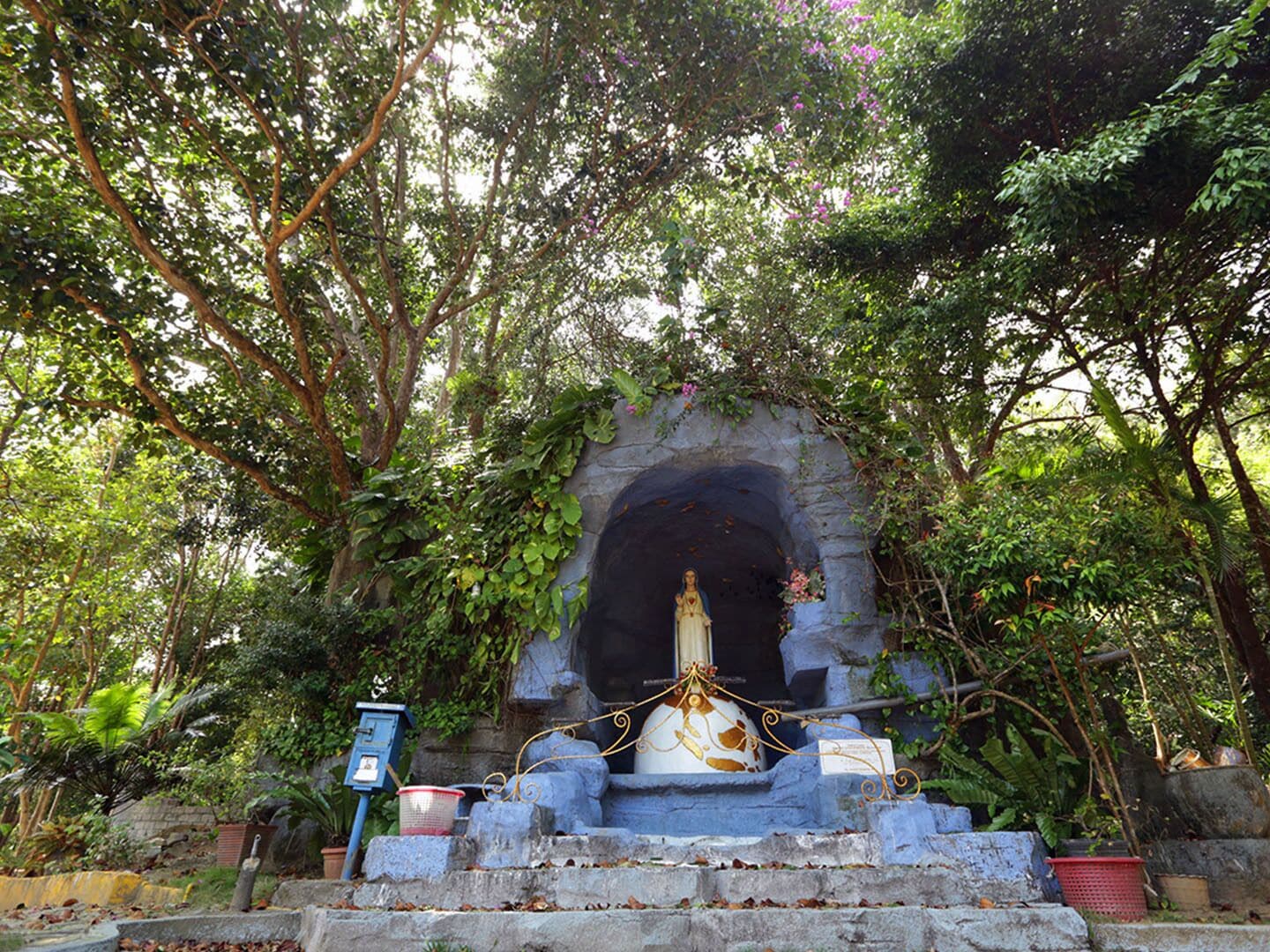 Eine Kapelle mit einer Statue, umgeben von Steinpodesten und Grünpflanzen.