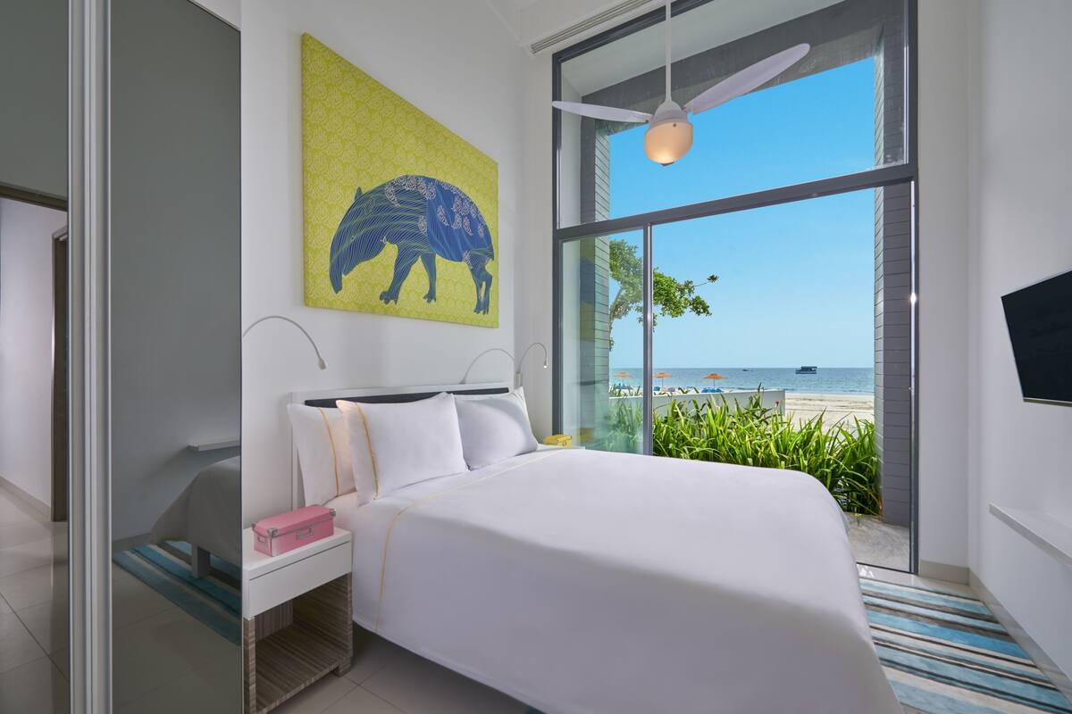 A modern hotel room with a large bed, a nightstand, a painting of a horse, and a view of the beach.