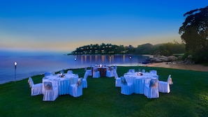 A lakeside outdoor dining setup with round tables and chairs, surrounded by a grassy area and a view of a calm lake and distant shoreline. - Cassia Bintan, part of Banyan Group (Bintan)