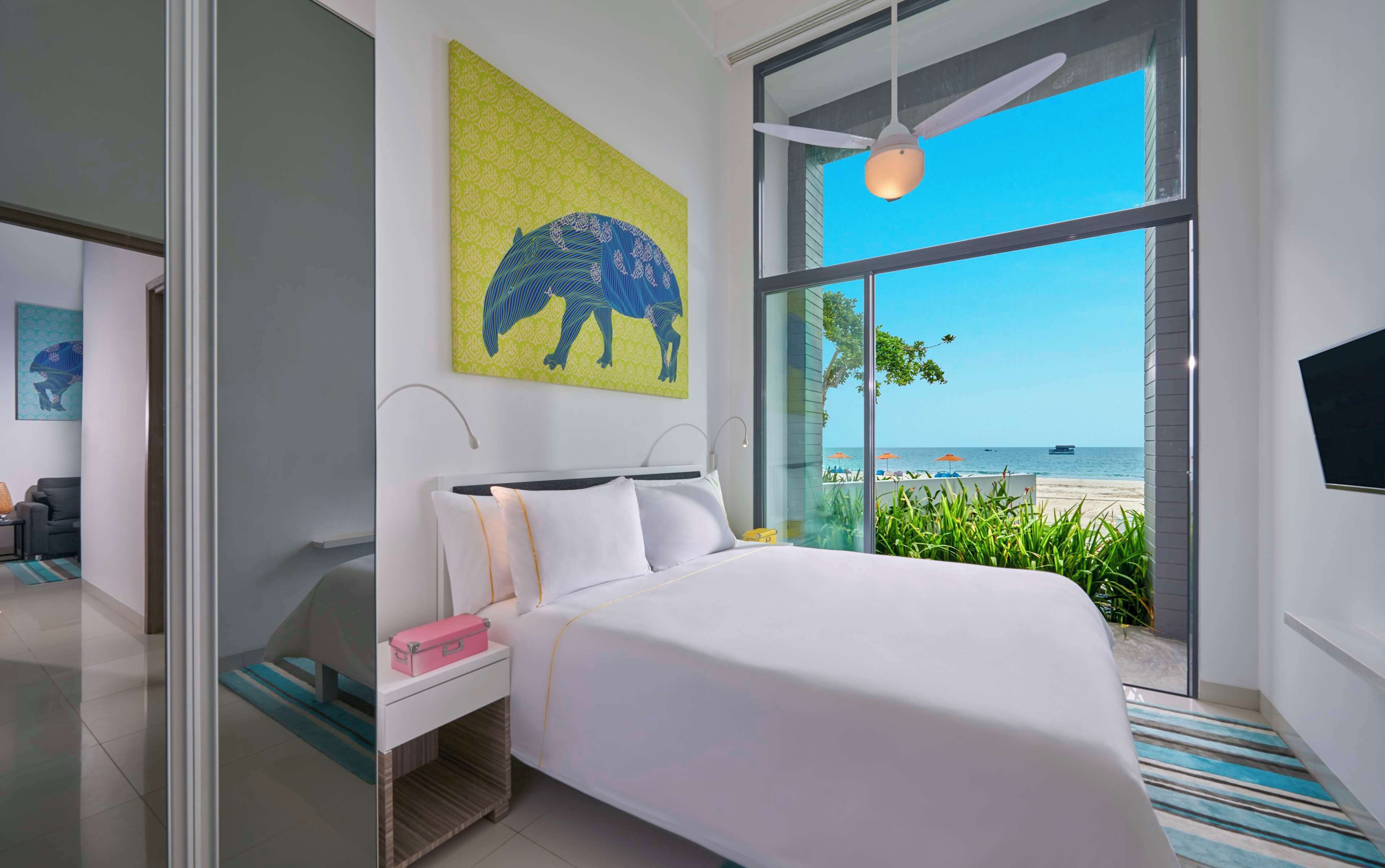 A modern hotel room with a large bed, a nightstand, a painting, and a view of the beach.
