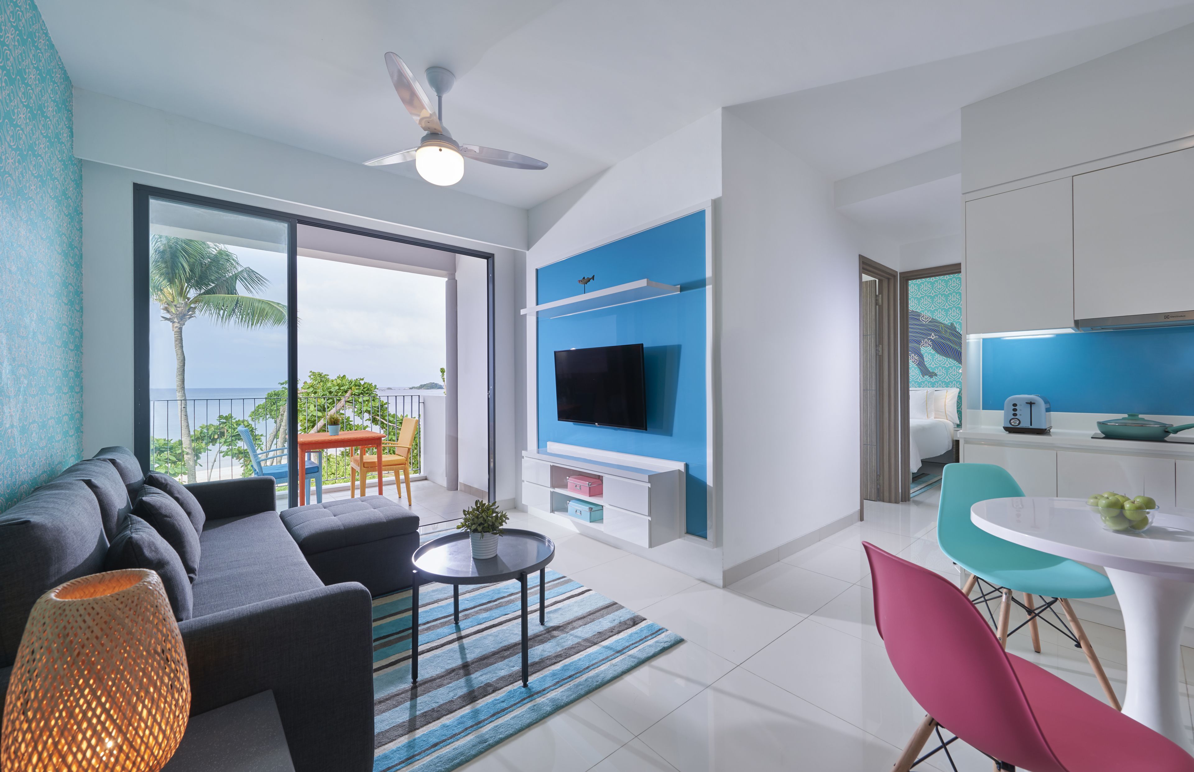 A modern living room with a sofa, coffee table, and TV. There is a balcony with a view of the ocean.