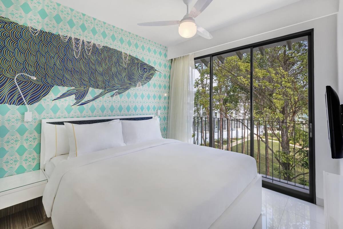 A bedroom with a bed, a ceiling fan, a large window with a view of trees, and a wall with a geometric pattern.