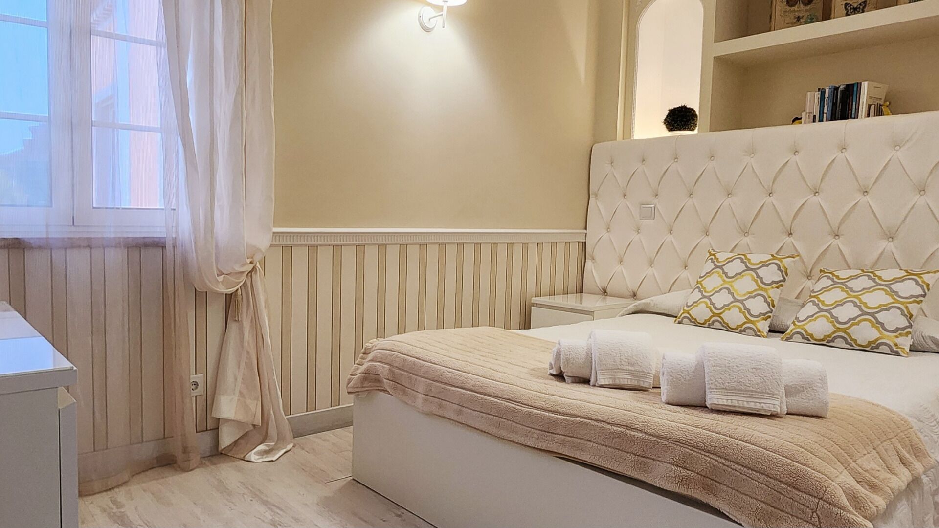 Une chambre à coucher avec une tête de lit capitonnée, une table de chevet, une lampe de chevet et une fenêtre avec des rideaux.