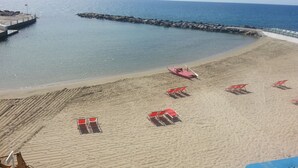 Une plage bordée de rangées de chaises de plage et de parasols rouges, une digue rocheuse et une vue dégagée sur l’océan.