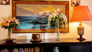 Une bibliothèque remplie de livres, un vase avec des fleurs, un tableau encadré représentant un paysage marin et une lampe de table.