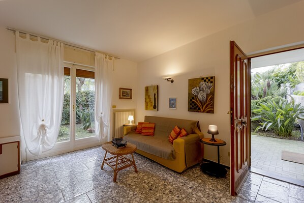 A living room with a sofa, a small table, and a chair. There is a door leading to a garden.